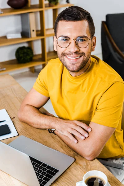 Homme souriant assis dans des lunettes près d'un ordinateur portable et une tasse de café — Photo de stock