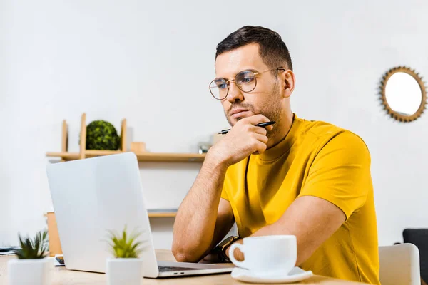 Задумчивый человек сидит в очках и смотрит на ноутбук возле чашки кофе — стоковое фото