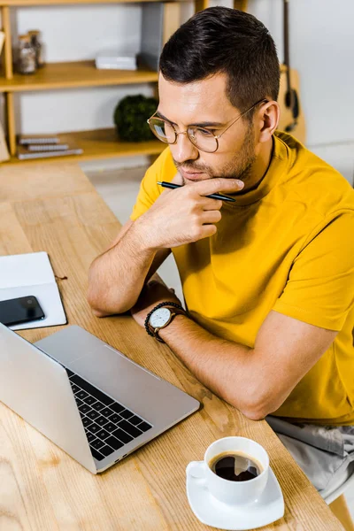 Homem pensativo sentado em óculos e olhando para laptop perto de xícara de café — Fotografia de Stock