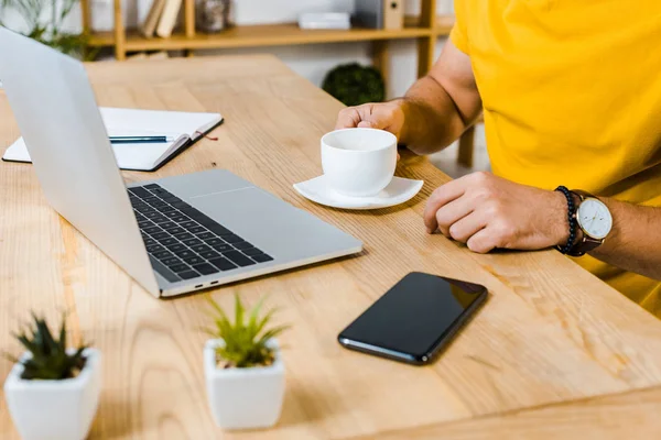 Cropped view of man holding cup of coffee near laptop and smartphone — Stock Photo