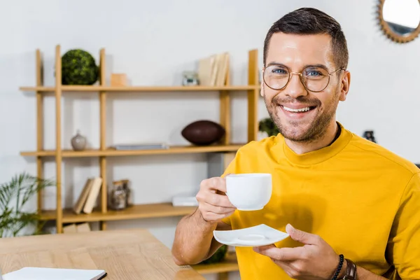 Sonriente hombre en vasos sosteniendo taza con café en casa - foto de stock