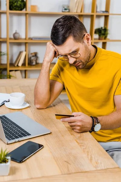 Uomo triste in occhiali guardando la carta di credito a casa — Foto stock