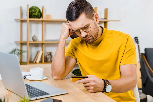 Upset man in glasses looking at credit card at home — Stock Photo