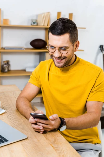 Homem bonito sorrindo enquanto olha para o smartphone em casa — Fotografia de Stock