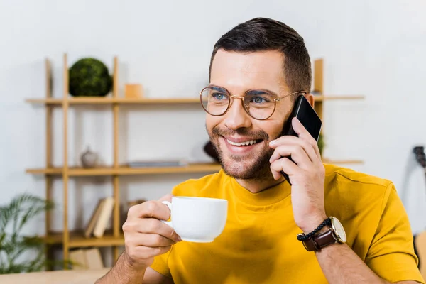 Bell'uomo in occhiali che parla sullo smartphone mentre tiene in mano una tazza di caffè — Foto stock