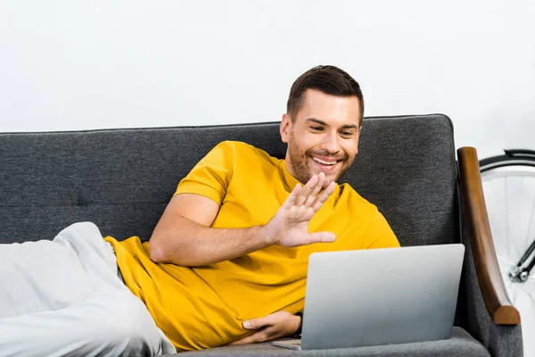Handsome man lying on sofa and waving to say hello while having video call — Stock Photo