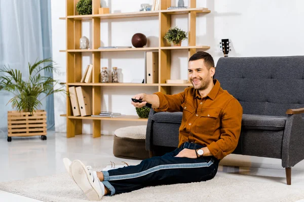 Homme joyeux tenant télécommande et assis sur le sol dans le salon moderne — Photo de stock