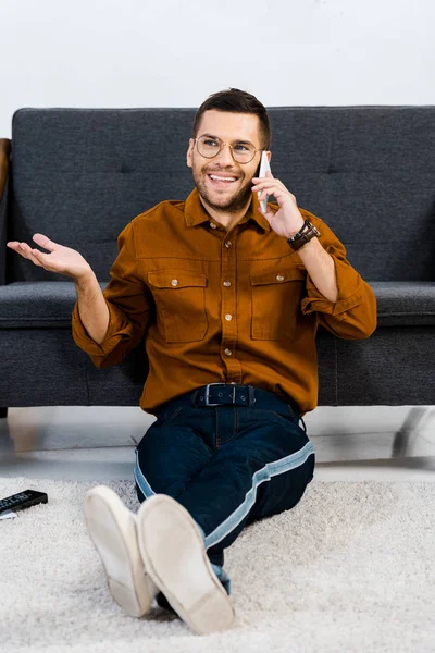 Cheerful man sitting on carpet and talking on smartphone — Stock Photo