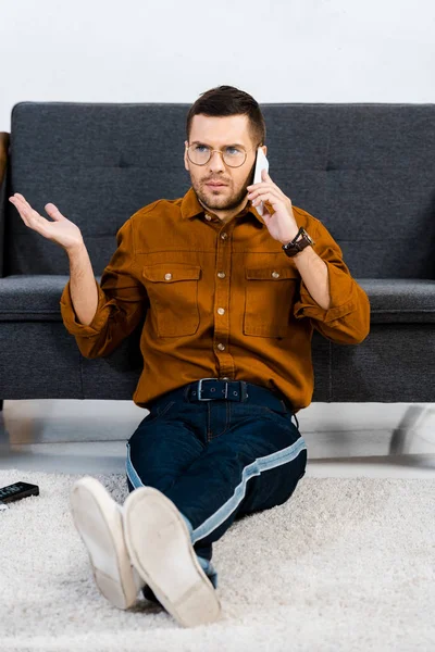 Dissatisfied man sitting on carpet and talking on smartphone — Stock Photo