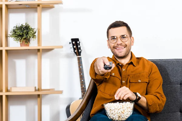 Cheerful man sitting on sofa with popcorn in bowl and holding remote control — Stock Photo