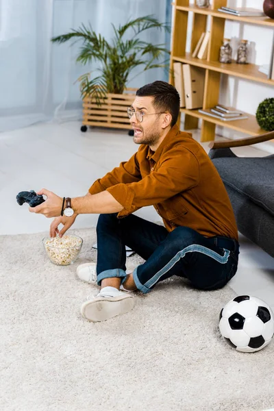 Emotional man sitting on carpet near popcorn while playing video game — Stock Photo
