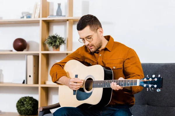 Красивый мужчина в очках играет на акустической гитаре в гостиной — стоковое фото