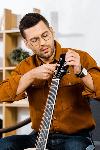 Bel homme à lunettes accordant guitare acoustique à la maison — Photo de stock