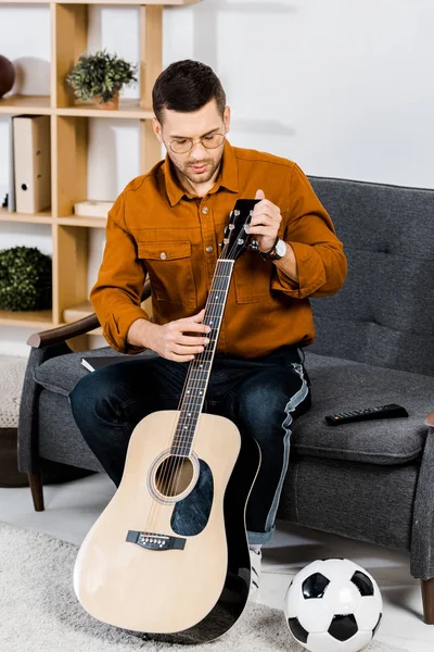 Homem bonito em óculos sentado no sofá e afinando guitarra acústica em casa — Fotografia de Stock