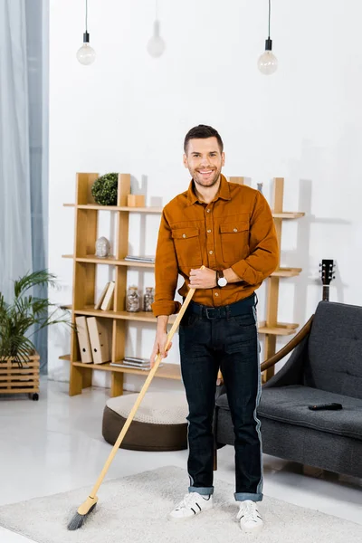 Uomo allegro in piedi con scopa in soggiorno moderno — Foto stock