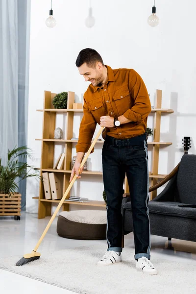 Hombre guapo alfombra de limpieza con escoba en la sala de estar moderna - foto de stock
