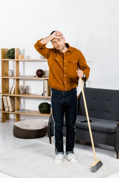 Homme fatigué debout avec balai dans le salon moderne — Photo de stock