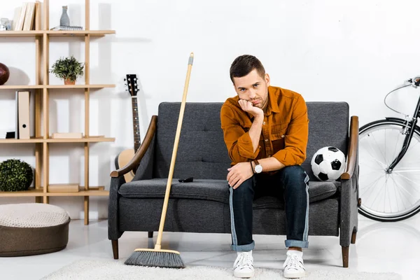 Hombre triste sentado en el sofá con escoba y bola en la sala de estar moderna - foto de stock
