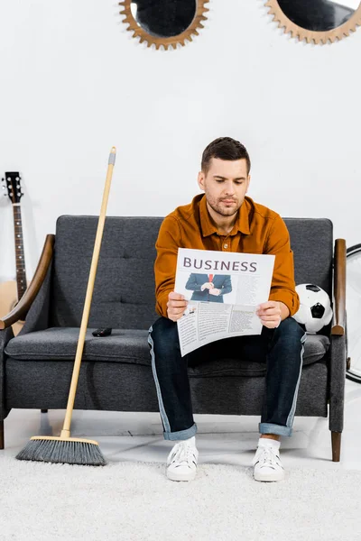 Schöner Mann sitzt auf Sofa neben Besen und liest Wirtschaftszeitung — Stockfoto