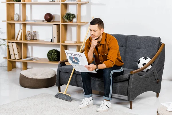 Homem sério sentado no sofá perto da vassoura e lendo jornal de negócios — Fotografia de Stock