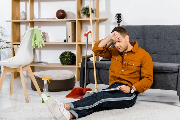 Homem bonito sentado no chão perto de equipamentos de limpeza na sala de estar moderna e jornal de leitura — Fotografia de Stock