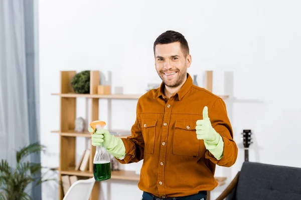 Hombre sonriente en la sala de estar moderna haciendo gesto pulgar hacia arriba y sosteniendo la botella - foto de stock