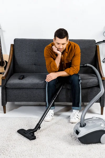 Upset man in modern living room sitting on sofa and looking at hoover — Stock Photo