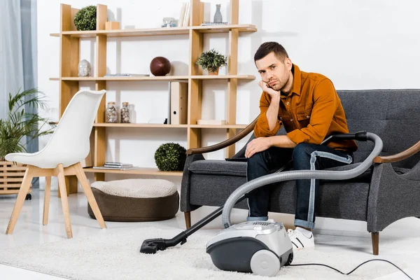 Upset man in modern living room sitting on sofa near hoover and looking at camera — Stock Photo