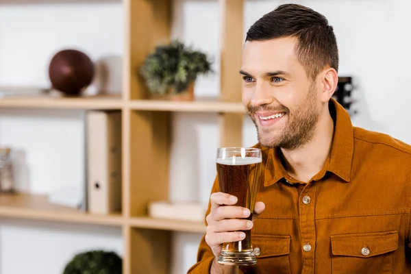 Hombre guapo en la sala de estar moderna sonriendo y sosteniendo un vaso de cerveza - foto de stock