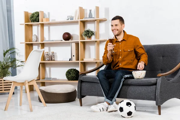 Schöner Mann im modernen Wohnzimmer auf dem Sofa sitzend, während er ein Glas Bier in der Hand hält und Popcorn isst — Stockfoto