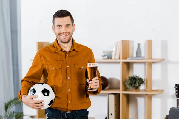 Bell'uomo in piedi nel moderno soggiorno mentre tiene palla da calcio e bicchiere di birra — Foto stock