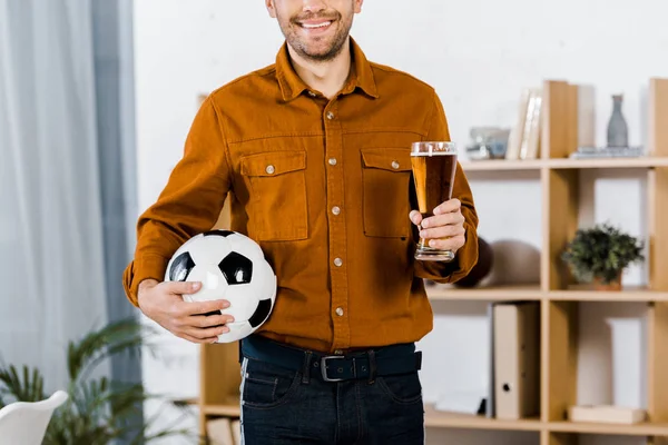 Vista cortada do homem de pé na moderna sala de estar segurando bola de futebol e copo de cerveja — Fotografia de Stock