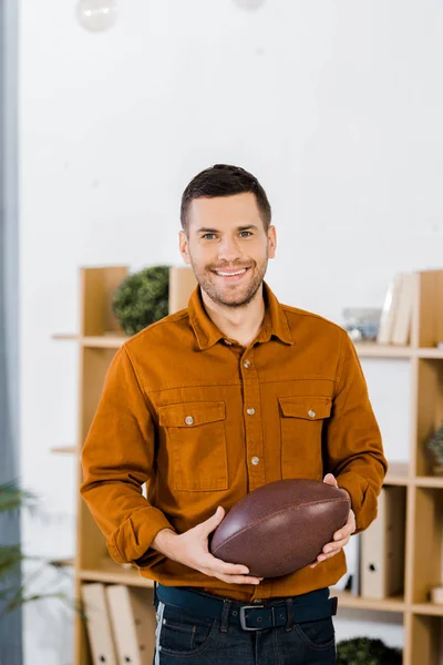 Homem bonito de pé na sala de estar moderna e segurando bola de futebol americano — Fotografia de Stock