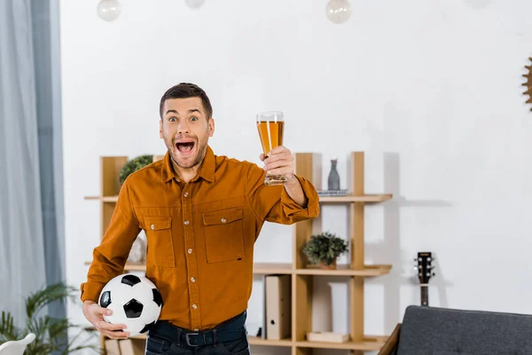 Bell'uomo in salotto moderno urlando mentre teneva palla da calcio e bicchiere di birra — Foto stock