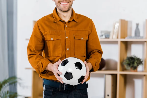 Vista ritagliata dell'uomo in piedi in salotto moderno con palla da calcio in mano — Foto stock