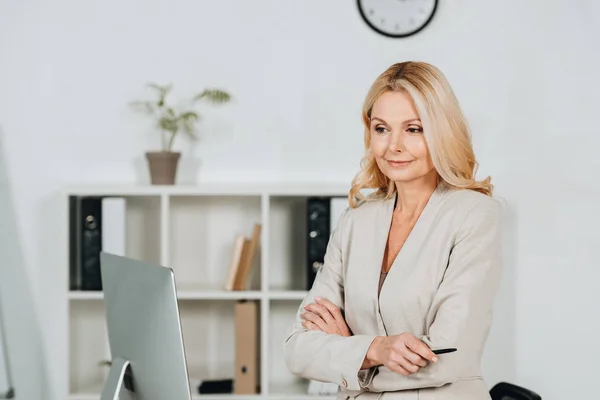 Beautiful mature businesswoman standing with crossed arms and looking at desktop computer in office — Stock Photo