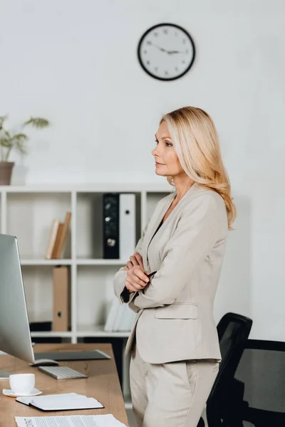 Side view of professional mature businesswoman standing with crossed arms and looking away in office — Stock Photo