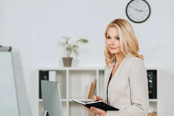 Задумчивая зрелая деловая женщина держит ноутбук и смотрит в сторону в офисе — стоковое фото