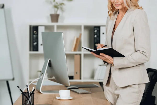 Schnappschuss einer lächelnden Geschäftsfrau, die am Arbeitsplatz steht und in Notizblock schreibt — Stockfoto