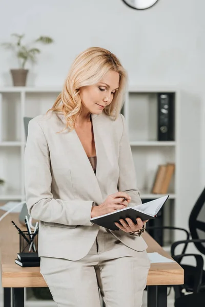 Focused mature businesswoman sitting on table and holding notepad in office — Stock Photo