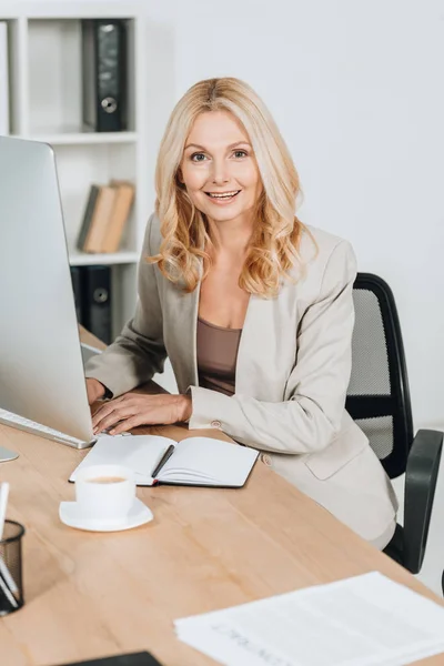 Alegre madura mujer de negocios usando computadora de escritorio y sonriendo a la cámara en la oficina - foto de stock
