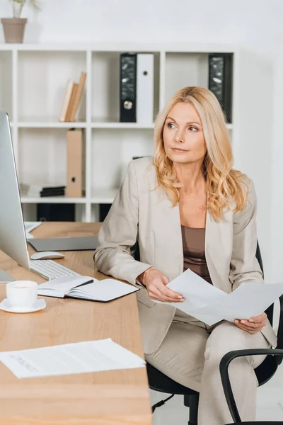 Focalizzata donna d'affari bionda in possesso di carte e guardando il computer desktop mentre seduto sul posto di lavoro — Foto stock