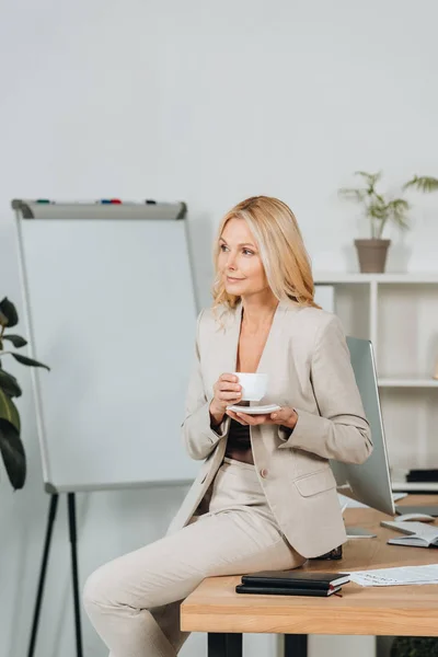 Красива усміхнена бізнес-леді тримає чашку кави і сидить на столі в офісі — стокове фото