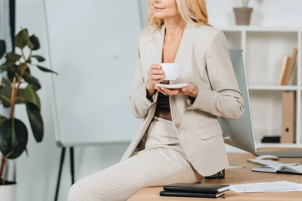 Tiro recortado de sorridente empresária segurando xícara de café e sentado na mesa no escritório — Fotografia de Stock
