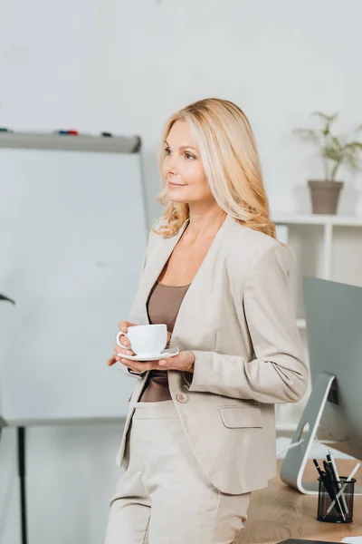 Красива усміхнена бізнес-леді тримає чашку кави і дивиться в офіс — стокове фото