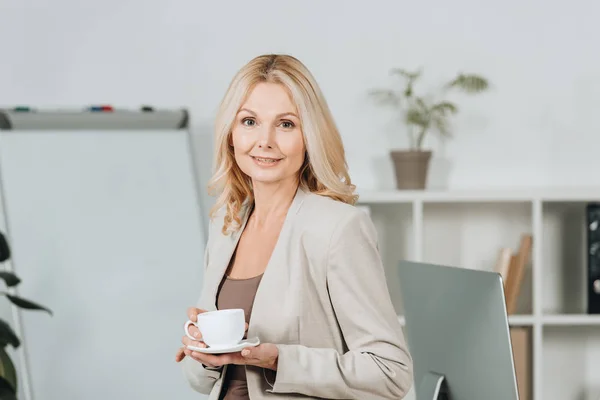 Hermosa mujer de negocios sosteniendo la taza de café y sonriendo a la cámara en la oficina - foto de stock