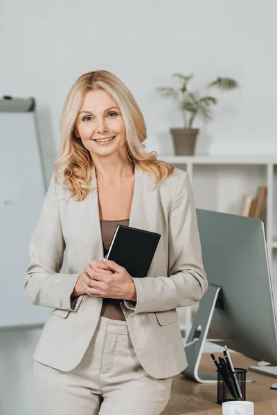 Beautiful mature businesswoman holding notebook and smiling at camera in office — Stock Photo