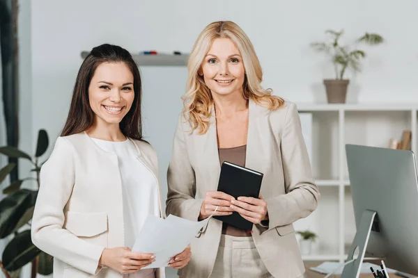 Hermosas mujeres de negocios profesionales con papeles y cuaderno de pie juntos y sonriendo a la cámara en la oficina - foto de stock