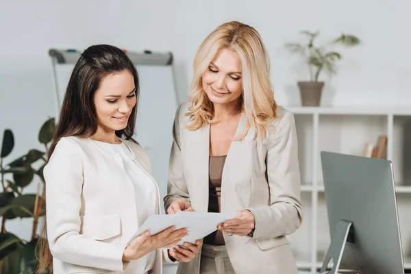 Schön lächelnde Geschäftsfrauen, die zusammen stehen und im Büro Papiere diskutieren — Stockfoto