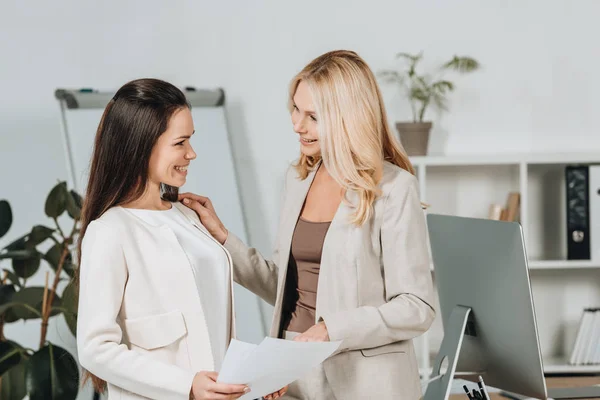 Lächelnde reife Geschäftsfrau mit Blick auf die schöne junge Kollegin, die Papiere im Büro hält — Stockfoto